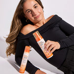A woman with long brown hair reclines, smiling and holding an orange Ultra Sheer Body Mist Sunscreen spray bottle. Next to her stands a Giovanni Cosmetics 2chic® ULTRA-VOLUME FOAM STYLING MOUSSE.
