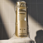 A gold bottle of Giovanni Cosmetics SMOOTHING CASTOR OIL SHAMPOO stands upright on a soap-sud-covered white ledge, with a tiled wall in the background.