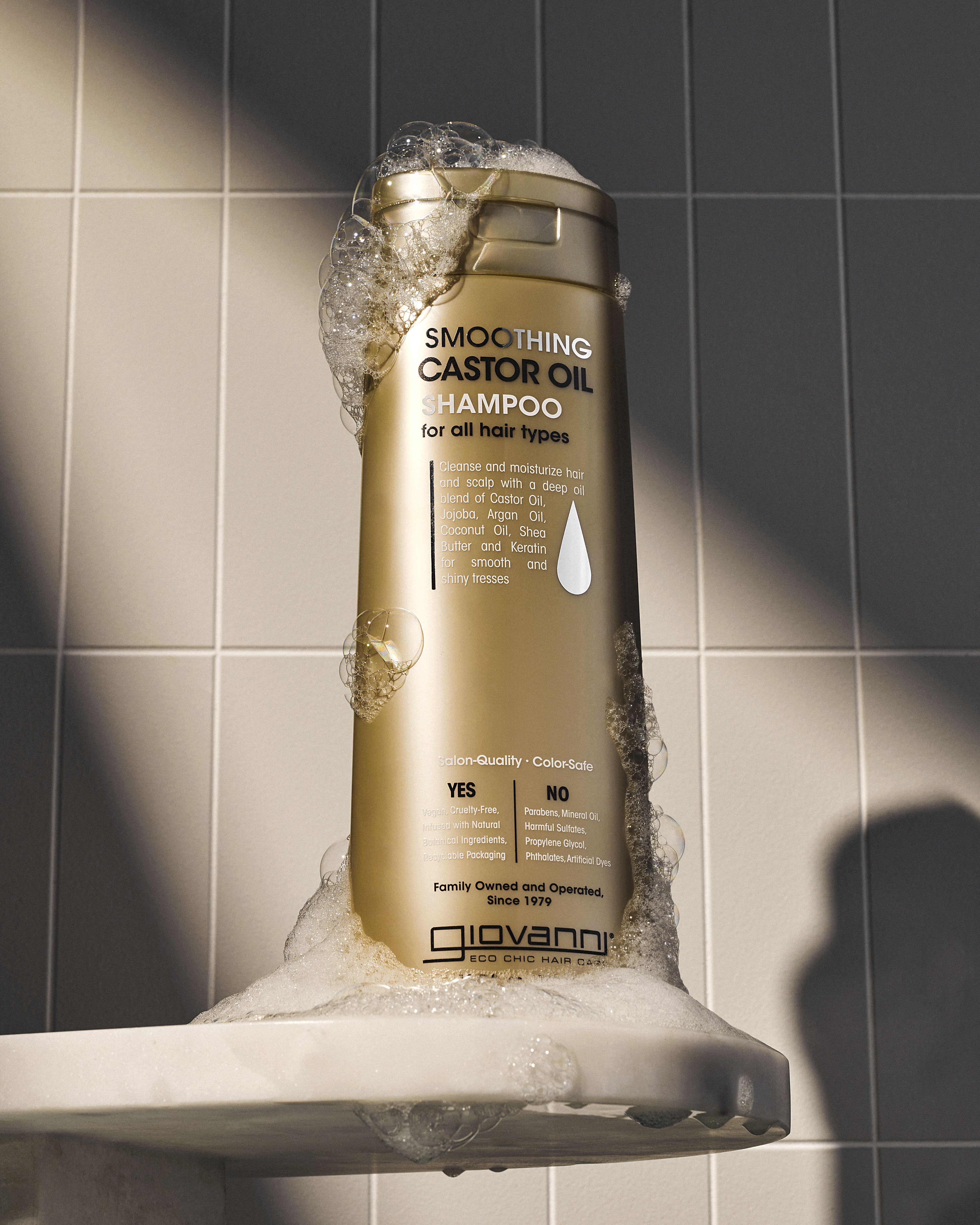 A gold bottle of Giovanni Cosmetics SMOOTHING CASTOR OIL SHAMPOO stands upright on a soap-sud-covered white ledge, with a tiled wall in the background.