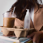 Person holding a carton with an iced coffee, a bottle of Giovanni's Root 66 Max Volume Directional Hair Root Lifting Spray, and a magazine.