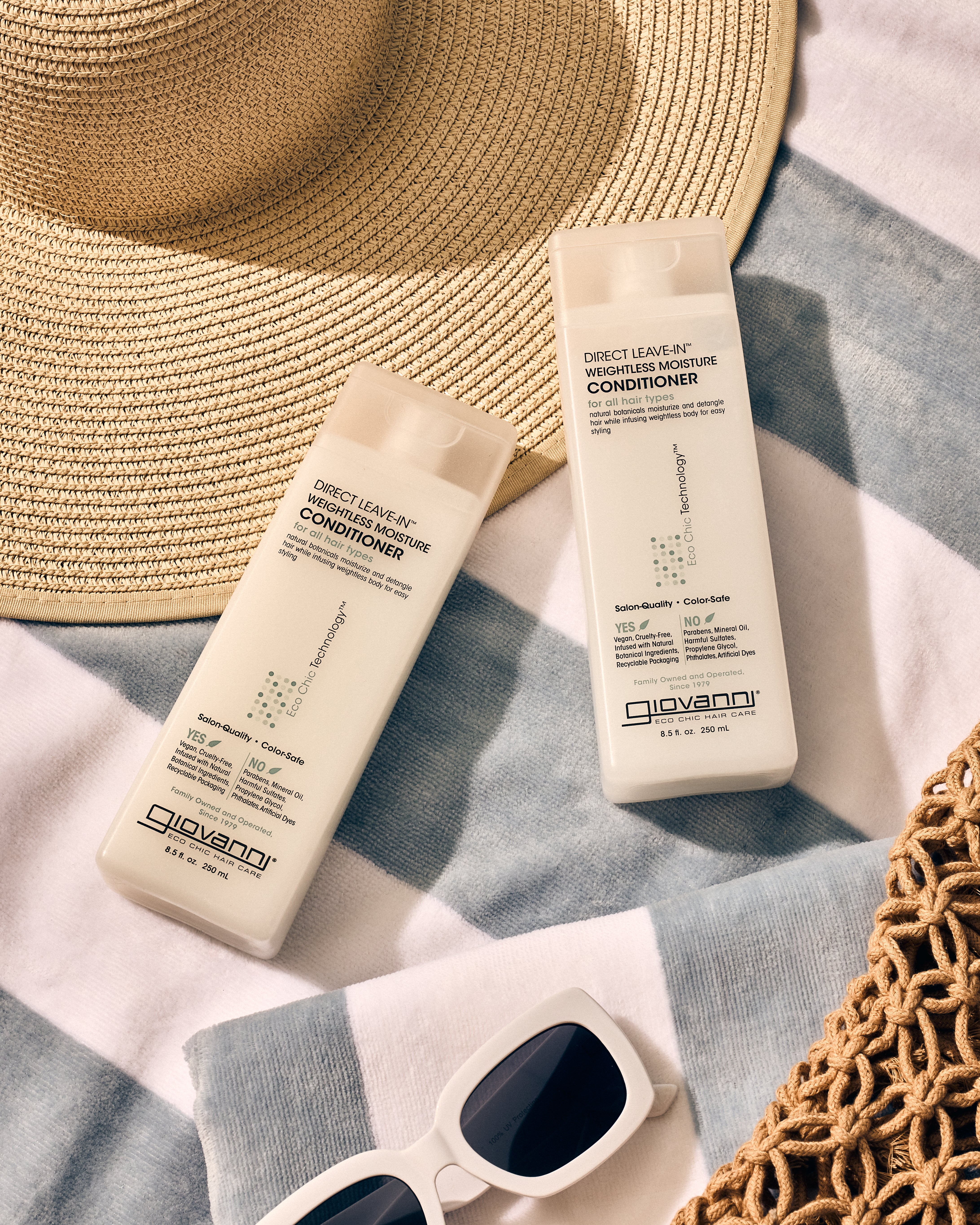 Two bottles of Giovanni Cosmetics DIRECT LEAVE-IN™ WEIGHTLESS MOISTURE CONDITIONER sit on a striped towel beside a sun hat, white sunglasses, and bag, highlighting weightless moisture in a relaxed, summery scene.