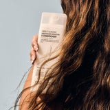 A person with long, wavy brown hair holds a translucent bottle of Giovanni Cosmetics DIRECT LEAVE-IN™ WEIGHTLESS MOISTURE CONDITIONER (2 Sizes) against their shoulder, set against a neutral light background.
