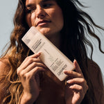 A woman with long wavy hair holds a bottle of Giovanni Cosmetics DIRECT LEAVE-IN™ WEIGHTLESS MOISTURE CONDITIONER close to her face, highlighting its weightless moisture benefits while looking away from the camera against a neutral background.