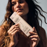 A woman with long wavy hair holds a bottle of Giovanni Cosmetics DIRECT LEAVE-IN™ WEIGHTLESS MOISTURE CONDITIONER close to her face, highlighting its weightless moisture benefits while looking away from the camera against a neutral background.