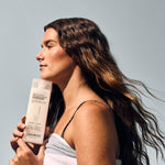 A woman with long, wavy brown hair holds Giovanni Cosmetics’ DIRECT LEAVE-IN™ WEIGHTLESS MOISTURE CONDITIONER—2 Sizes—near her face. Wrapped in a white towel, she radiates natural beauty against a light grey background.