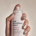 A hand with shiny silver nail polish holds a white can of Giovanni Cosmetics HAIR TEXTURIZING SPRAY for styling and finishing, mist visible against a neutral background—perfect for boosting hair volume.