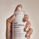 A hand with shiny silver nail polish holds a white can of Giovanni Cosmetics HAIR TEXTURIZING SPRAY for styling and finishing, mist visible against a neutral background—perfect for boosting hair volume.