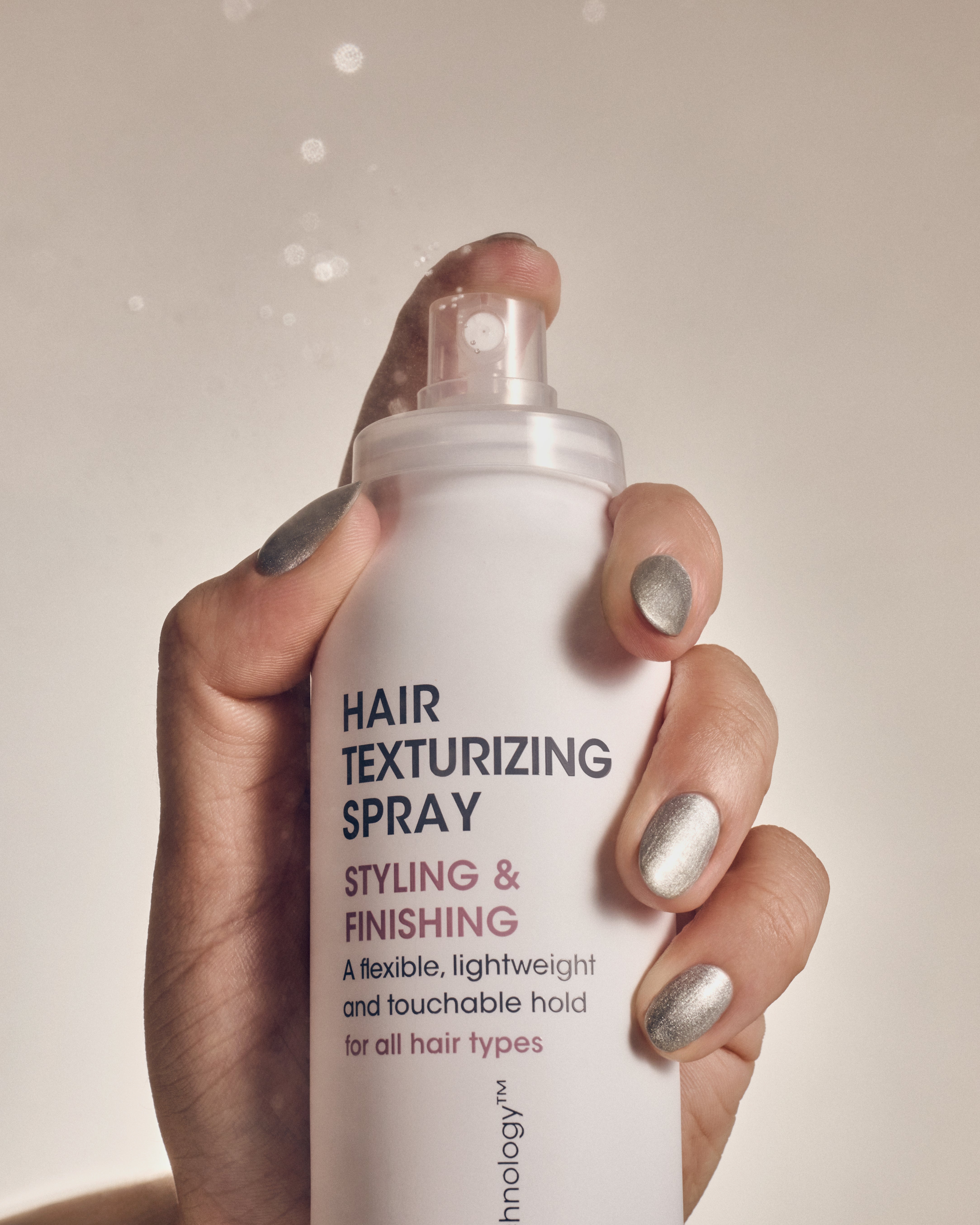 A hand with shiny silver nail polish holds a white can of Giovanni Cosmetics HAIR TEXTURIZING SPRAY for styling and finishing, mist visible against a neutral background—perfect for boosting hair volume.