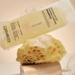 A bottle of Giovanni Cosmetics 50:50 Balanced Hydrating-Clarifying Shampoo is shown tilted over a yellow loofah sponge with soap bubbles around it on a warm beige background.