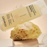 A bottle of Giovanni Cosmetics 50:50 Balanced Hydrating-Clarifying Shampoo is shown tilted over a yellow loofah sponge with soap bubbles around it on a warm beige background.