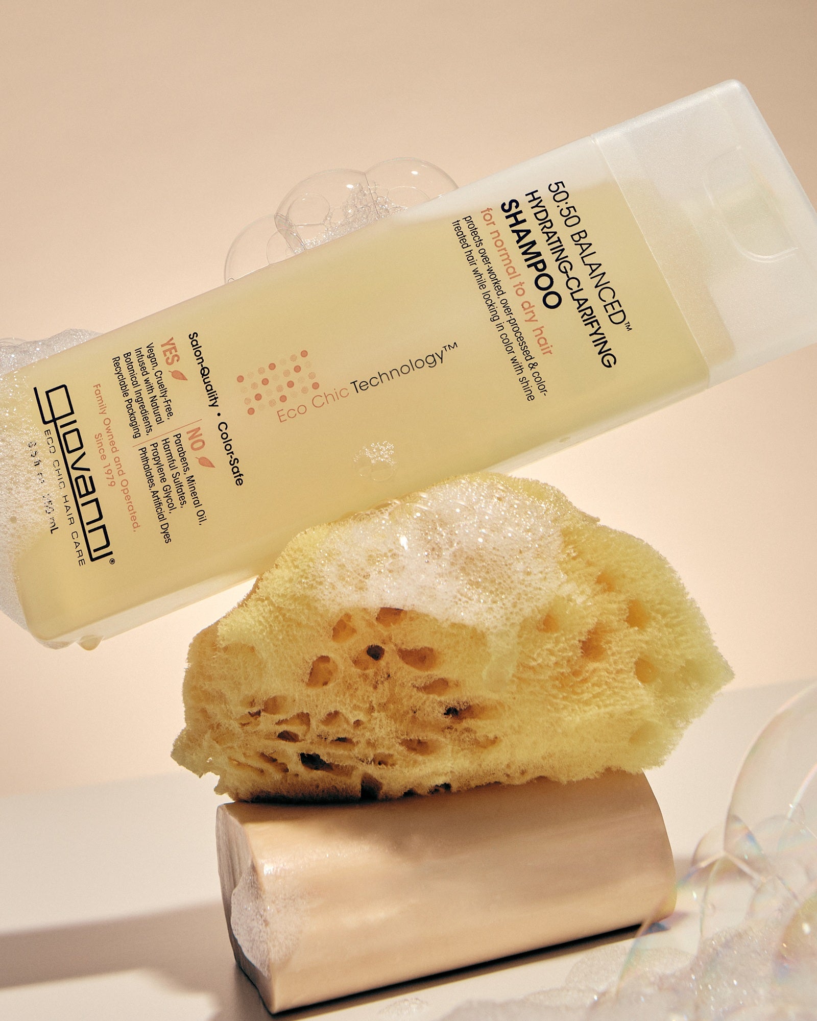 A bottle of Giovanni Cosmetics 50:50 Balanced Hydrating-Clarifying Shampoo is shown tilted over a yellow loofah sponge with soap bubbles around it on a warm beige background.