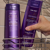 Two bottles of Curl Habit Curl Defining Shampoo and Curl Habit Curl Defining No-Foam Conditioning Shampoo with a hand holding one bottle, removing it from a shelf within a shower. 