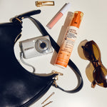 A black leather crescent shoulder bag with gold hardware, accompanied by a silver digital camera, a pink lip gloss tube, two gold bobby pins, tortoiseshell sunglasses and a bottle of Ultra Volume Leave-In Conditioning & Styling Elixir on a white background.