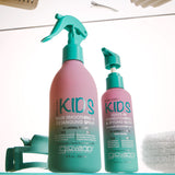 Two Giovanni Kids hair care bottles on a teal shelf: a larger pink spray bottle labeled Hair Smoothing & Detangling Spray and a smaller pink bottle labeled Conditioner & Styling Balm, with teal pump tops. A comb, hair clips, and a towel are partially visible around them.