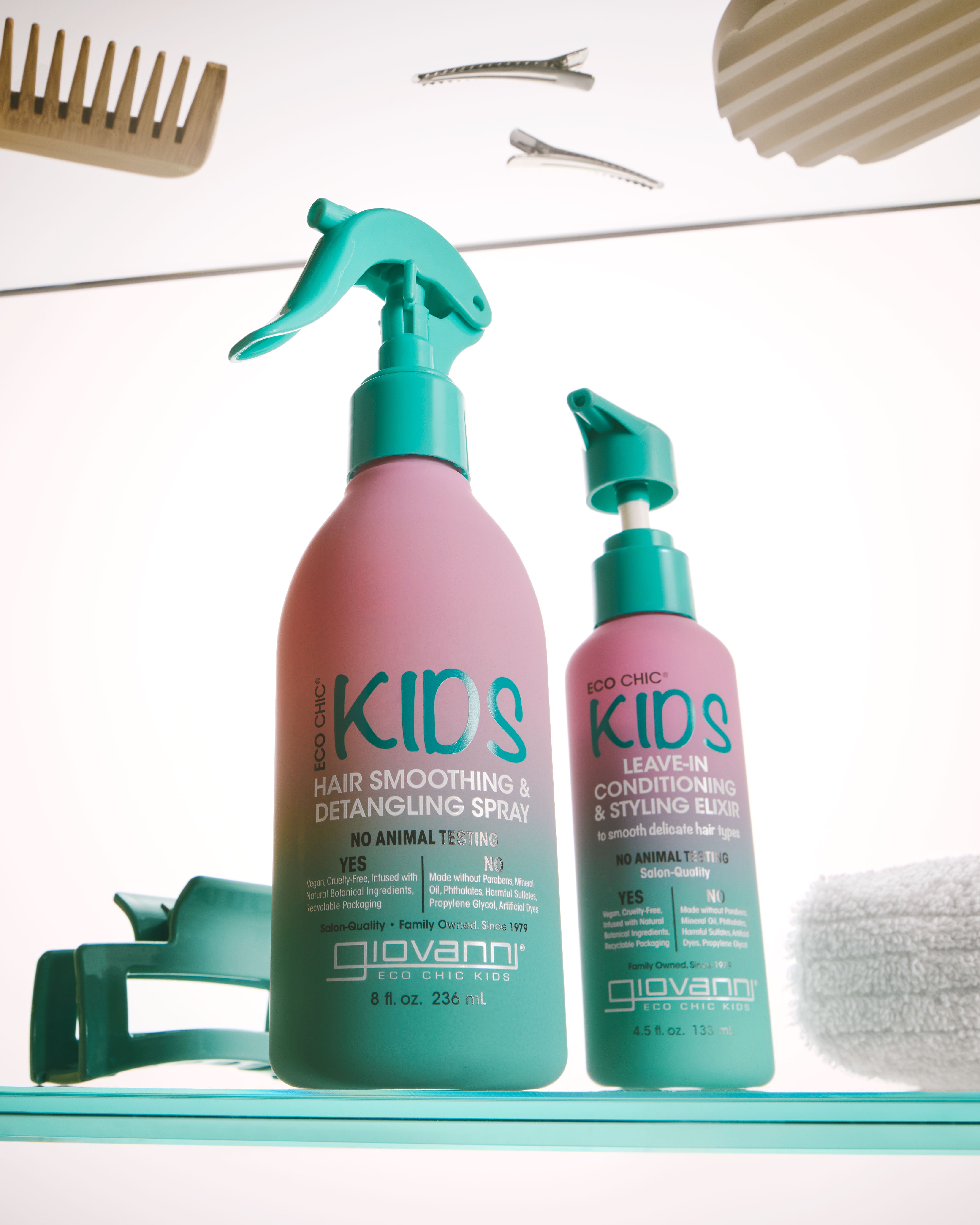 Two Giovanni Kids hair care bottles on a teal shelf: a larger pink spray bottle labeled Hair Smoothing & Detangling Spray and a smaller pink bottle labeled Conditioner & Styling Balm, with teal pump tops. A comb, hair clips, and a towel are partially visible around them.
