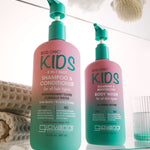 Two Giovanni Eco Chic Kids hair care bottles on a glass shelf: a larger pink-and-teal shampoo and conditioner bottle, and a smaller matching body wash bottle, with bath accessories softly blurred in the background.