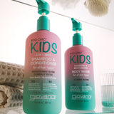 Two Giovanni Eco Chic Kids hair care bottles on a glass shelf: a larger pink-and-teal shampoo and conditioner bottle, and a smaller matching body wash bottle, with bath accessories softly blurred in the background.