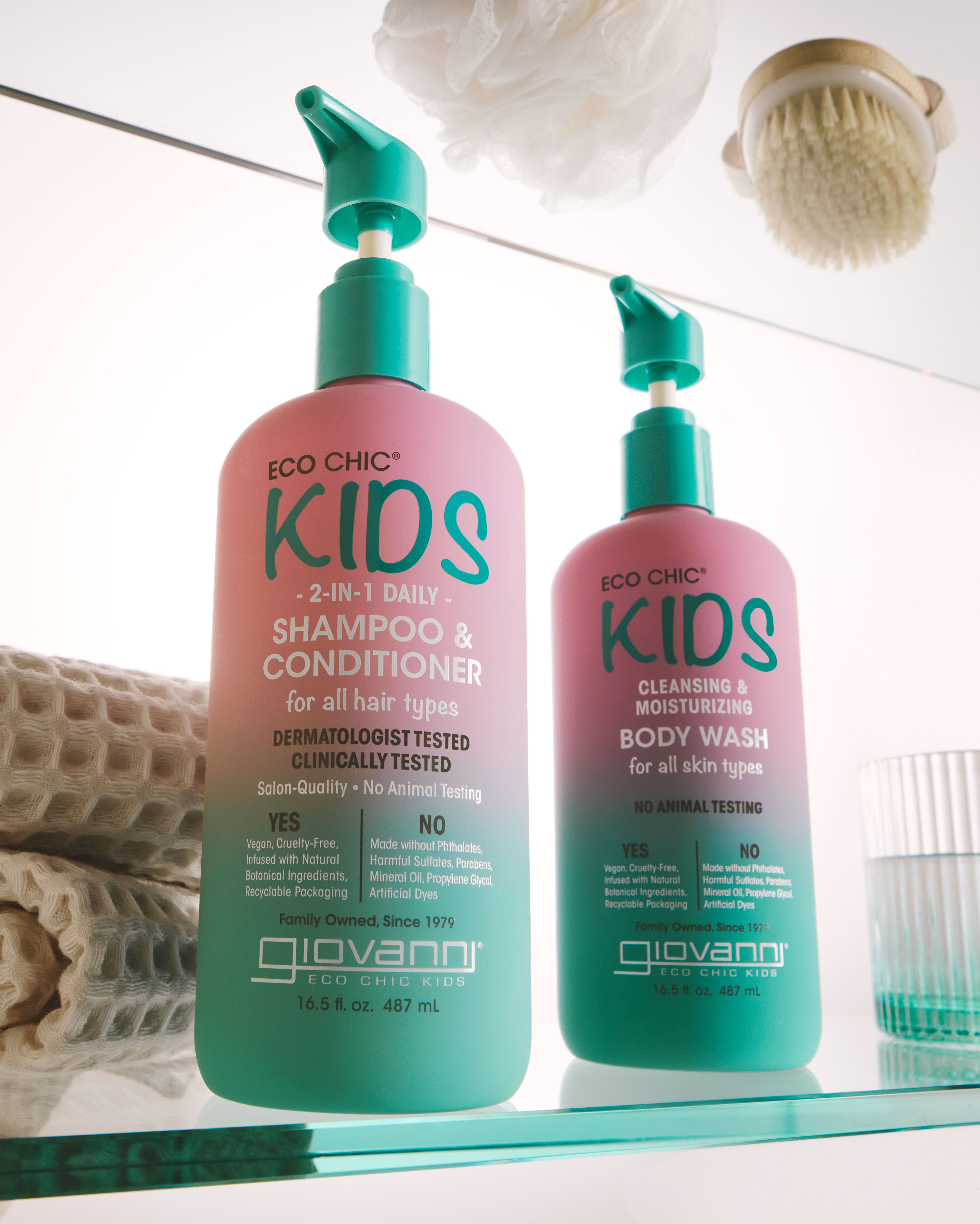 Two Giovanni Eco Chic Kids hair care bottles on a glass shelf: a larger pink-and-teal shampoo and conditioner bottle, and a smaller matching body wash bottle, with bath accessories softly blurred in the background.