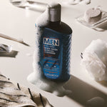 Giovanni Men body wash and facial cleanser bottle surrounded by white foam on a bathroom surface, with a towel, soap sponge, glass, and sponge visible in a styled setting.