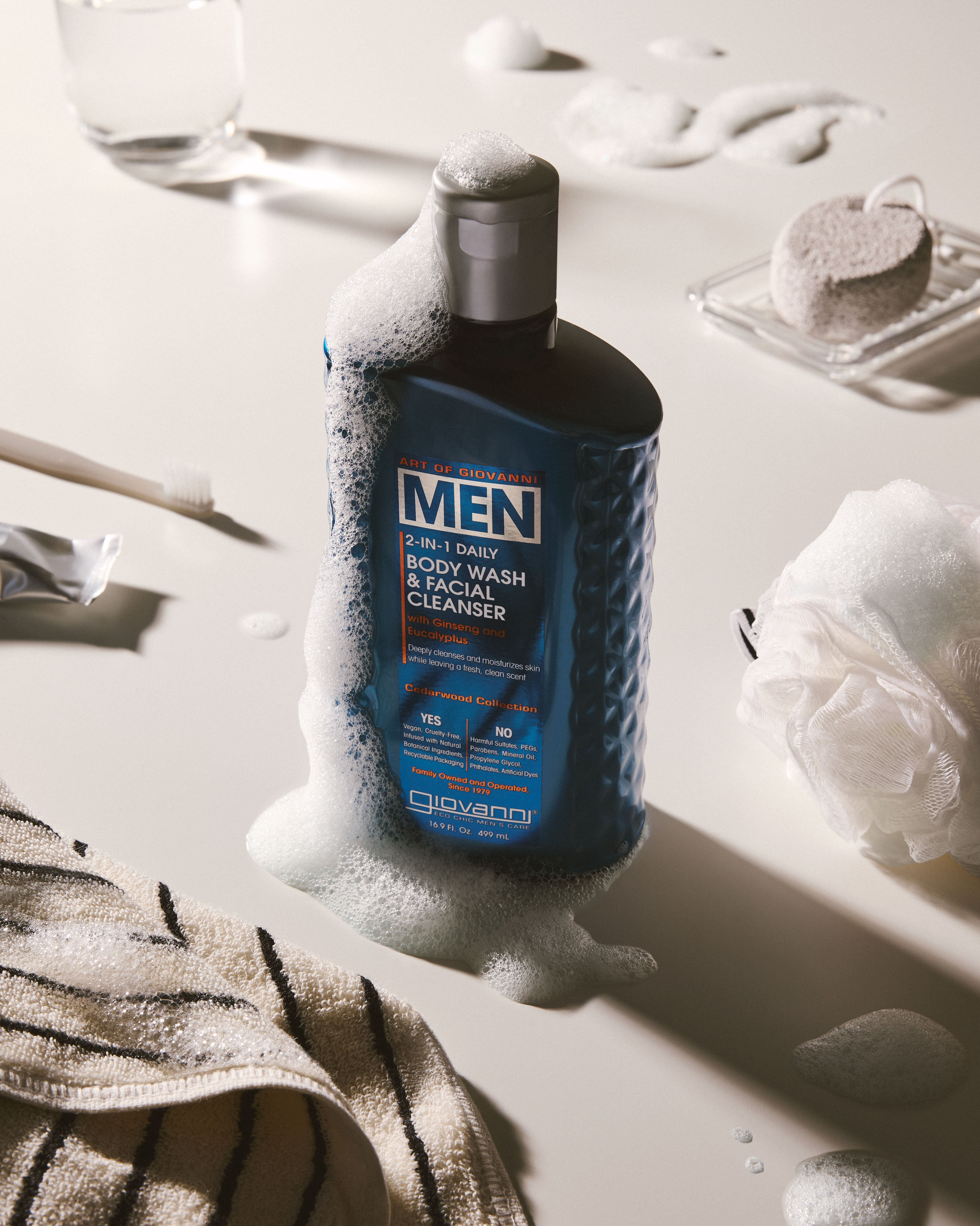 Giovanni Men body wash and facial cleanser bottle surrounded by white foam on a bathroom surface, with a towel, soap sponge, glass, and sponge visible in a styled setting.