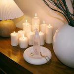 A frosted pump bottle with a white label and gold logo stands on a round white tray on a wooden table, styled with lit white candles, a pleated lamp, and a large white vase with dried branches.