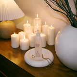 A frosted pump bottle with a white label and gold logo stands on a round white tray on a wooden table, styled with lit white candles, a pleated lamp, and a large white vase with dried branches.