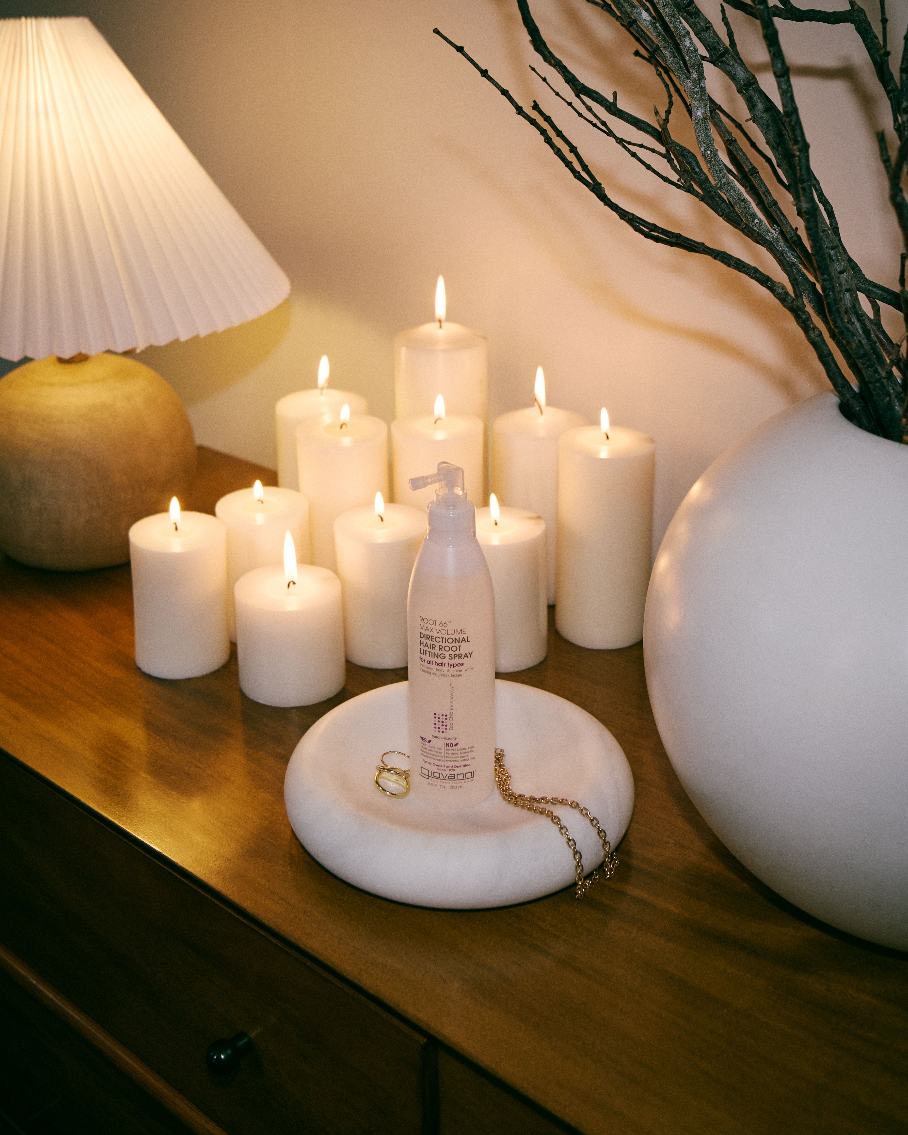 A frosted pump bottle with a white label and gold logo stands on a round white tray on a wooden table, styled with lit white candles, a pleated lamp, and a large white vase with dried branches.
