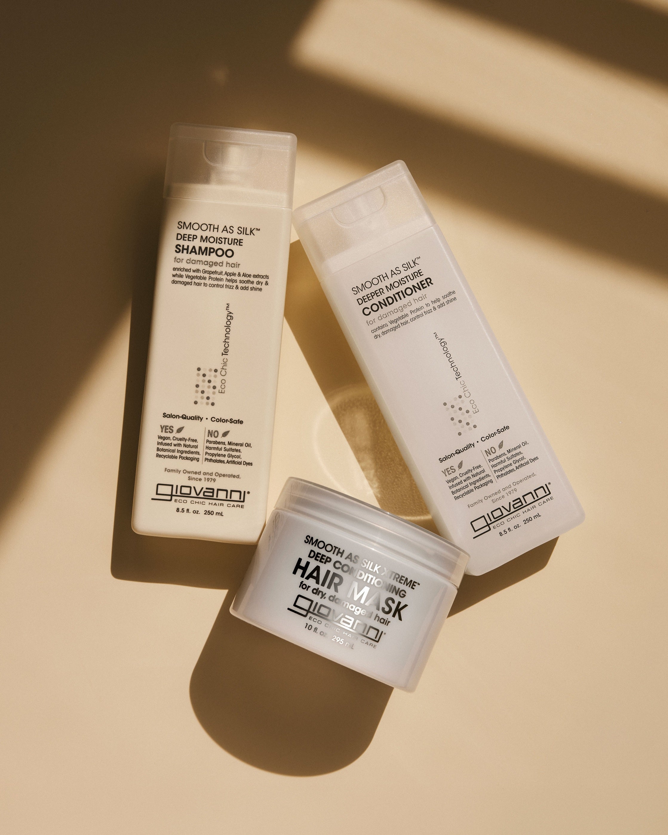 Three Giovanni Smooth As Silk haircare products in white packaging are arranged diagonally on a warm beige surface with soft diagonal shadows.