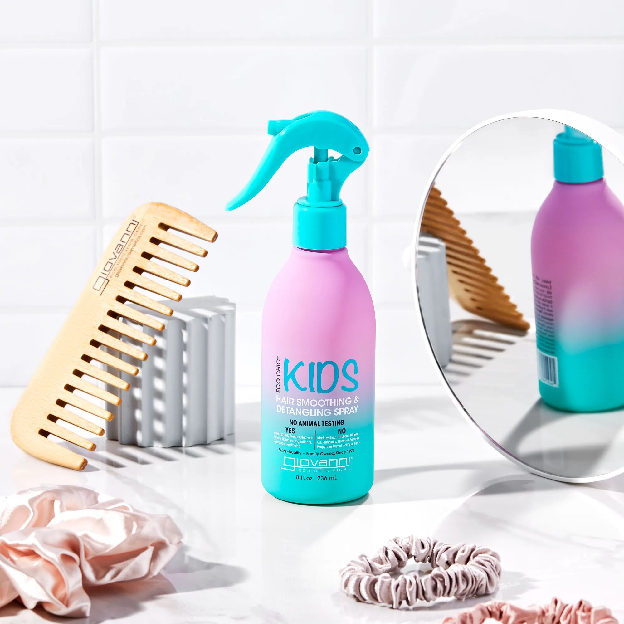 A bottle from the Giovanni Cosmetics KIDs Bundle stands on a counter alongside a wooden wide-tooth comb, scrunchies, and a round mirror, all set against a white tile background.