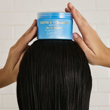 Person holding a jar of Giovanni Biotin & Collagen hair mask over long black hair against a white tiled wall.