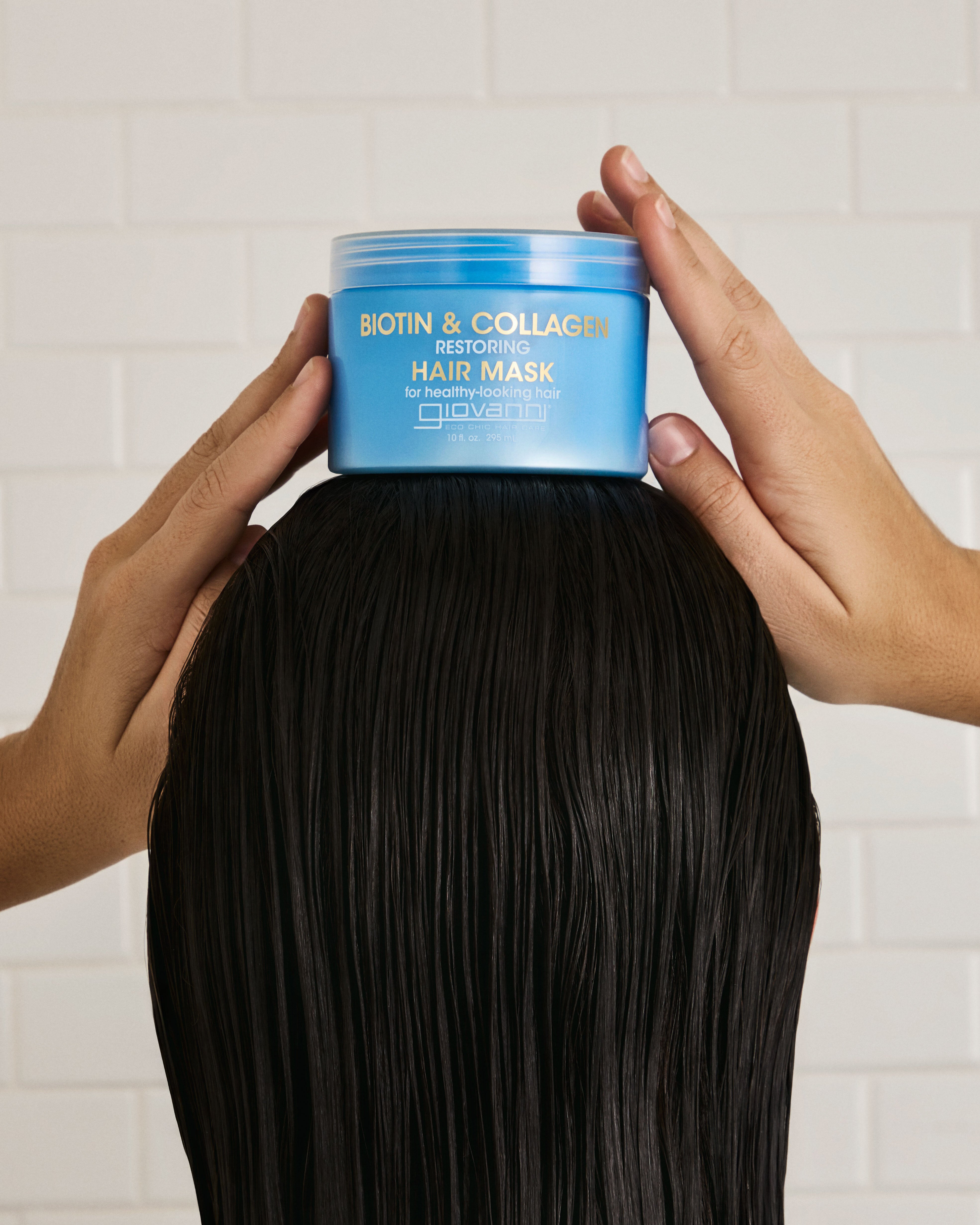 Person holding a jar of Giovanni Biotin & Collagen hair mask over long black hair against a white tiled wall.