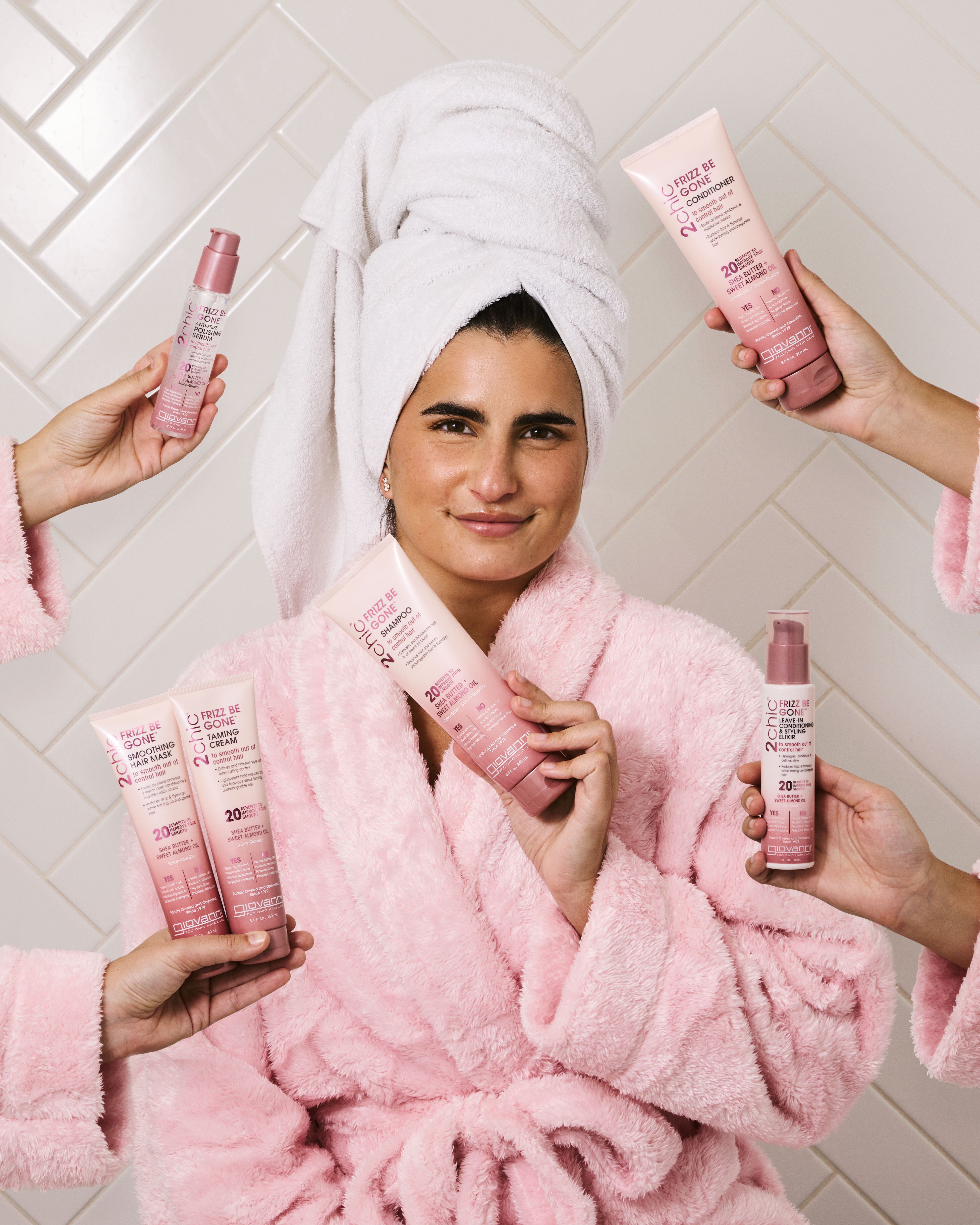 Woman in a pink robe holding a bottle of 
Giovanni 2Chic Frizz Be Gone Shampoo surrounded by hands holding other Frizz Be Gone products