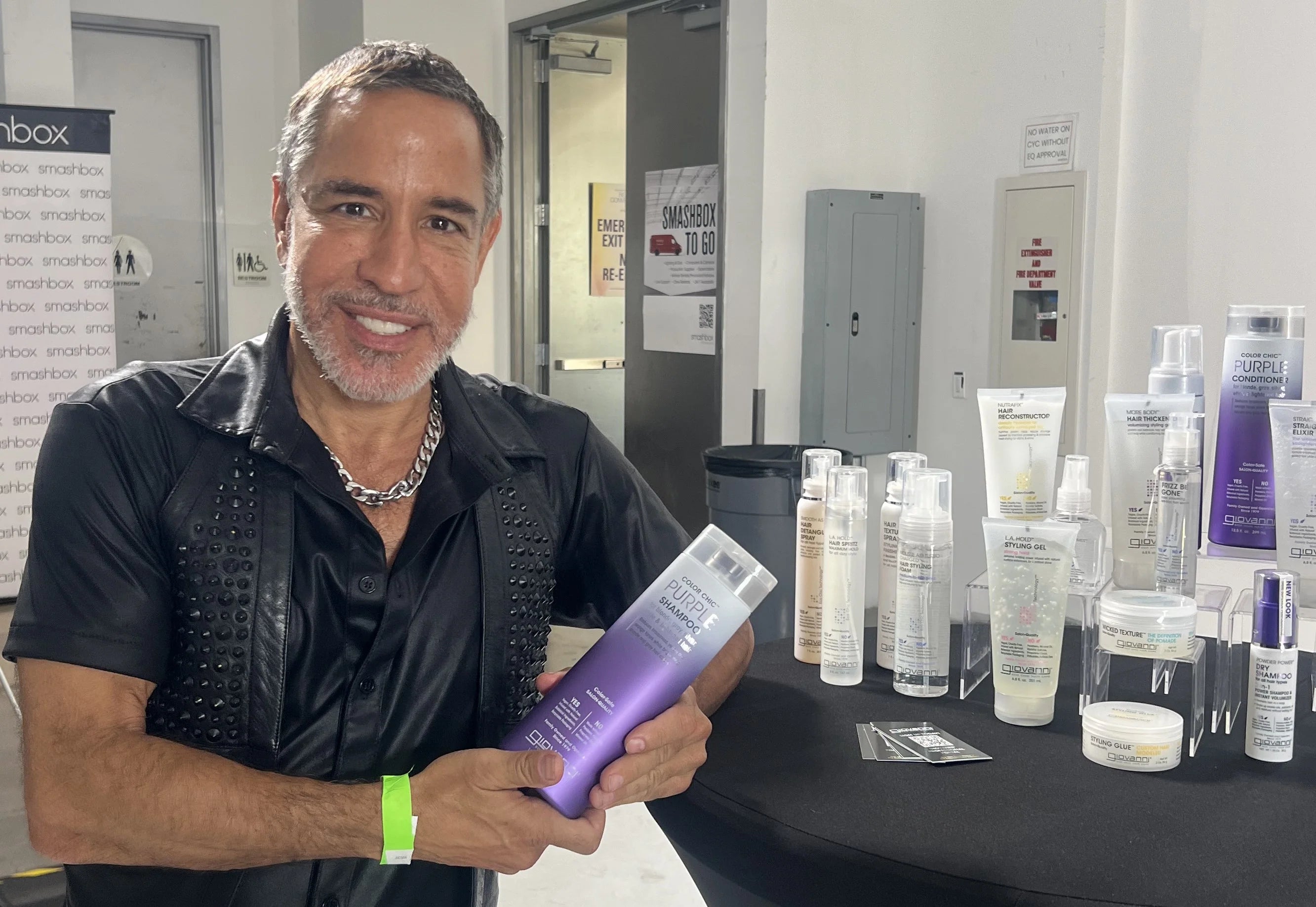 A person smiles and holds a bottle of Giovanni Cosmetics COLOR CHIC™ PURPLE SHAMPOO at a display table with various hair and beauty products arranged neatly in a well-lit indoor setting, ready to banish brassy tones for brighter blonde hair.