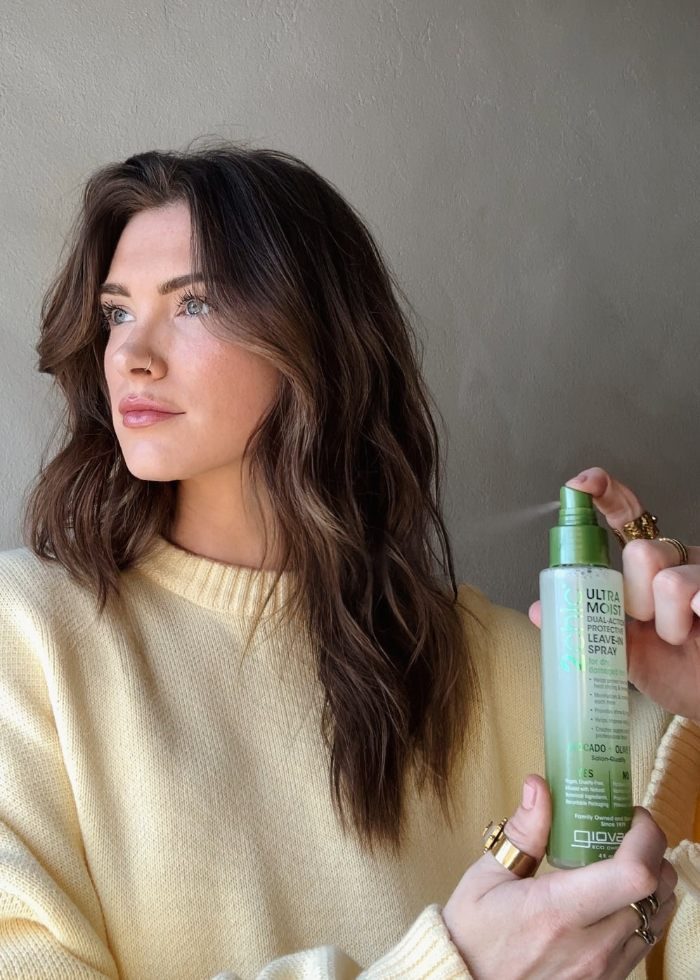 A woman with long brown wavy hair in a pale yellow sweater looks to the side as another hand holds Giovanni Cosmetics 2chic® ULTRA-MOIST DUAL-ACTION PROTECTIVE LEAVE-IN SPRAY with avocado and olive oil near her hair.