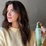 A woman with long brown wavy hair in a pale yellow sweater looks to the side as another hand holds Giovanni Cosmetics 2chic® ULTRA-MOIST DUAL-ACTION PROTECTIVE LEAVE-IN SPRAY with avocado and olive oil near her hair.