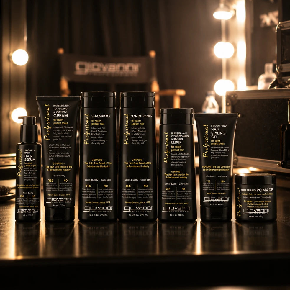 Bottles of Giovanni Professional Collection products on a dark reflective surface, including shampoo, conditioners, hair serum, hair styling gel, and pomade, with warm blurred lights in the background.